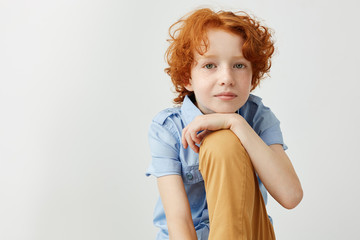 Wall Mural - Close up of beautiful ginger boy in blue shirt and yellow jeans holding hand on leg, looking in camera and gently smiling.