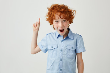 Wall Mural - Close up of funny ginger boy with freckles pointing apside on white background with opened mouth and silly face expression. Copy space