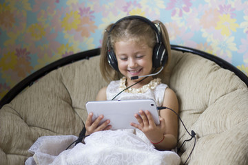 Wall Mural - little girl in a beautiful dress in headphones with a microphone sitting in a chair and looks at the tablet