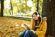 © ViDi Studio - A brown-haired smiling woman in yellow coat and jeans sitting and listening to music under a tree with a tablet in her hands and headphones in fall city park on a warm day. Autumn golden leaves.