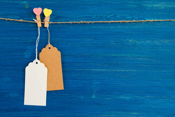 Two brown and white blank paper price tags or labels set and wooden pins decorated on hearts hanging on a rope on the blue wooden background.