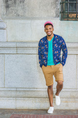 African American Man Casual Street Fashion in New York, wearing blue flower patterned jacket, green V neck T shirt, yellow brown shorts, white sneakers, pink cap, standing by vintage wall, smiling..