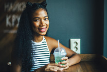 Beautiful Smiling Hipster Teenager Black Ecuadorian Woman Tasting Smoothie Or Juice In Modern Interior Cafe. Relaxing, Lifestyle And Leisure Concept. Fancy Hairstyle Like A Cat. Close Up.