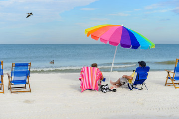 Wall Mural - elderly couple sitting on sun loungers with a colorful beach umbrella on silver sand  vacation concept