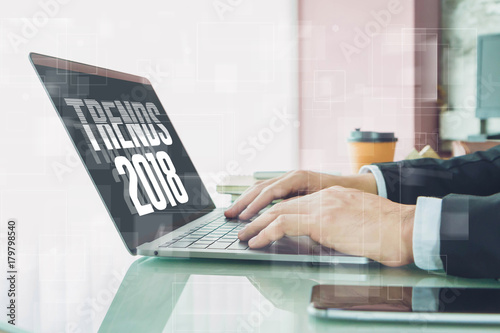 Fotografía Hand of Businessman typing " trends 2018" on laptop screen with smartphone and coffee cup in home office or co working space
