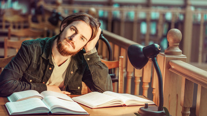 Portrait of thinking young bearded student man reading in a library hall on table with lot of books and lamp, indoor dusk time, education concept