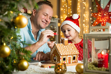 Father And Adorable Daughter In Red Hat Building Gingerbread House Together. Beautiful Decorated Room With Lights And Christmas Tree, Table With Candles And Lanterns. Happy Family Celebrating Holiday.
