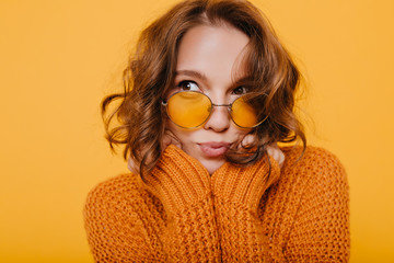 Close-up portrait of cute pensive girl with shiny curls looking away on yellow background. Indoor photo of romantic female model in glasses and knitted sweater thinking about something.