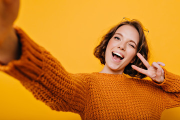 indoor portrait of blissful girl in knitted clothes making selfie and laughing on bright background.