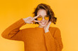© Look! - Joyful european girl with shiny curls laughing and showing peace sign on yellow background. Indoor portrait of beautiful lady in glasses and knitted clothes having fun in studio.