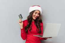 Pretty Cheerful Brunette Woman In Santa's Hat Holding Credit Card And Laptop While Shopping Online