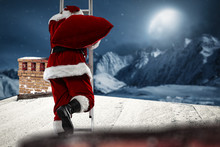 The Old Santa Claus Enters The Roof Of The House. Landscape Of The Mountains And The Big Moon. Visible Big Chimney On The Roof.