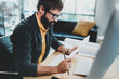© SFIO CRACHO - Closeup of stylish bearded designer in eye glasses working at sunny loft studio.Man using contemporary desktop computer and making notes in documents.Horizontal.Blurred background.
