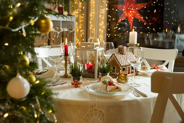 Beautiful served table with decorations, candles and lanterns. Little gingerbread house with glaze on white tablecloth. Living room decorated with lights and Christmas tree. Holiday setting