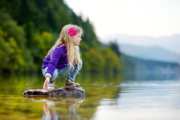 Wall Mural - Adorable girl playing by Hallstatter See lake in Austria on warm summer day. Cute child having fun splashing water and throwing stones into the lake.