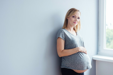 Young pregnant woman standing against gray wall, smiling. 