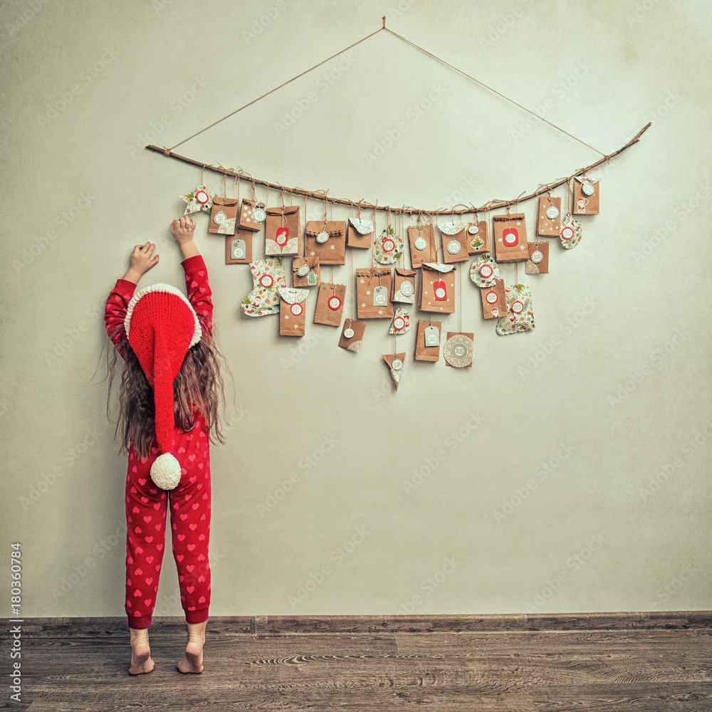 child in pajamas and Christmas cap stretches for advent calendar with small gifts - obrazy, fototapety, plakaty 