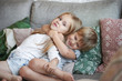 Sweet shot of two adorable little sisters hugging on sofa in living room: baby girl with short hair embracing her elder sister tight, holding hands around her neck, surrounded with decorative pilows