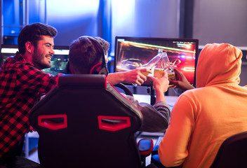 Canvas Print - Strong friendship. Back view of joyful stylish young guys are sitting at table while clinking of bottles of beer. They are enjoying video game on computer while spending time at home. Selective focus