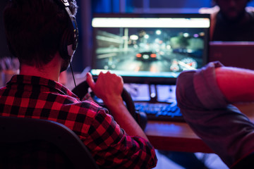 Back view of concentrated young gamer in headphones is playing car racing video game at home with steering wheel. He is looking at monitor of modern computer. Selective focus