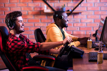 Wall Mural - Entertainments of real male. Optimistic stylish guys are spending time in computer club. Focus on joyful bearded gamer is playing car racing video game with steering wheel. African man in background