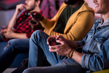 Adult entertainment. Close-up of hands with joystick of young man who is playing home video game while relaxing on couch and expressing gladness. His pleasant stylish friends are sitting in background