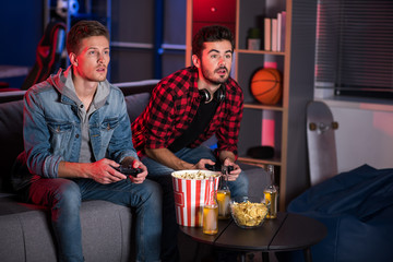 Wall Mural - Weekend entertainment. Involved trendy cute guys are sitting on couch and holding joystick while playing home video games. They are looking at screen absorbedly. Copy space in the right side