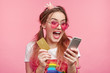 Headshot of fashionable young female looks crazy as verifies balance, happy to recieve large sum of money, uses smart phone with mobile banking application, keeps credit plastic card in hands
