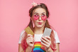 Horizontal portrait of female chick has dreamful expression, imagines new clothes, going do online shopping, holds modern cell phone and plastic card, isolated over pink background with copy space