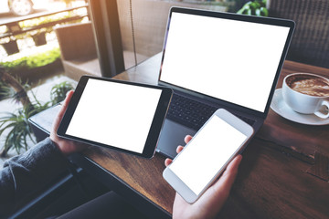 Mockup image of a businesswoman holding white mobile phone , black tablet and laptop with blank white screen on vintage wooden table with coffee cup in cafe
