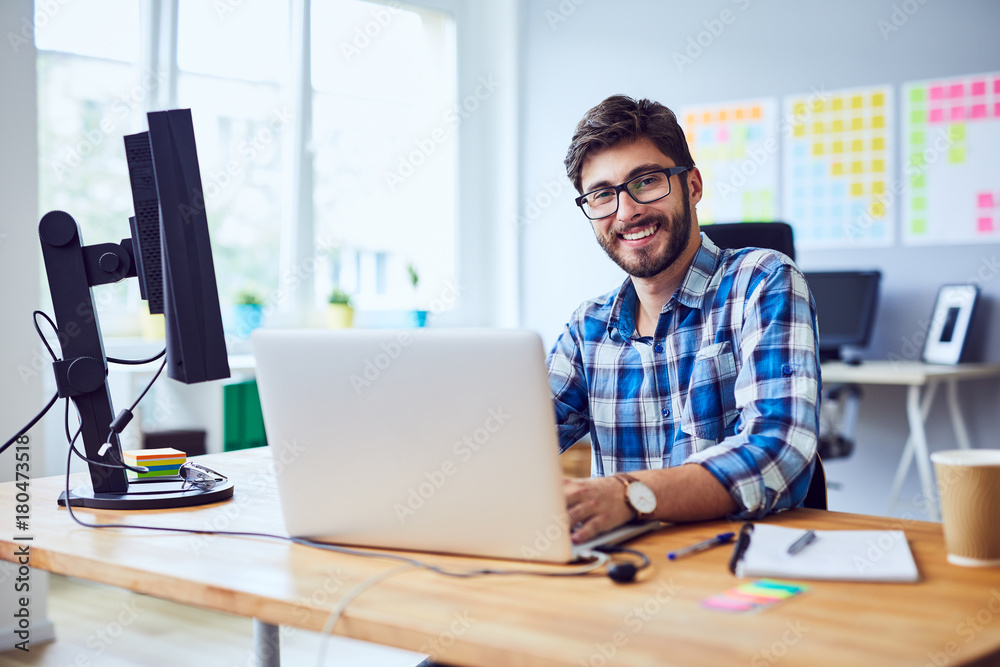 Cheerful young programmer looking at camera while working at his office - obrazy, fototapety, plakaty 