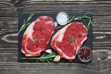 Raw beef steak with spices and rosemary on black slate board over wooden background