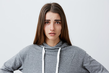 Close up portrait of young good-looking unhappy caucasian girl with dark long hair in casual grey hoodie crying, looking in camera with wet eyes after breakup with boyfriend. Negative emotions