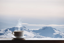 Cup Of Hot Drink With Steam On Wooden Desk And Snow Capped Mountain View In The Morning