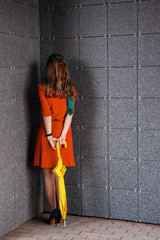 A young elegant girl posing in red dress with yellow umbrella. The girl is standing with her face to the corner.