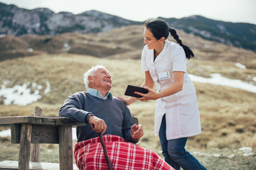 Wall Mural - Smiling caregiver nurse and  disabled senior patient using digital tablet outdoor