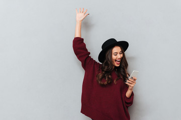 Wall Mural - Brunette woman in sweater and hat using smartphone and rejoice