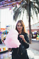 Wall Mural - Beautiful young woman or girl stands in middle of country fair carnival, enjoys holiday food, festival pink sugar candy cotton or floss, in front of attraction ride on summer break date or day out