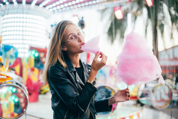 Wall Mural - Cute and pretty young girl or student, eats and poses with sugar pink candy cotton, wears leather jacket in line for attraction ride or rollercoaster on town festival, happy times in summer