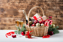 Two Glasses Of Champagne With Christmas Basket With Bottle, Gifts With Red Satin Ribbon, Candy Canes, Pine Cones, Golden Garlands On Dark Brown Wooden Background, New Year Celebration Concept