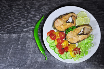 fresh fish, cut into steaks, fried in butter and seasoned with spices, served on a festive white plate with tomatoes, cucumbers, chilli peppers and leeks on a black table as background