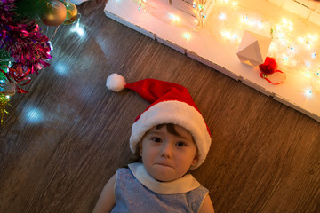 Funny girl in Santa hat laying on the floor near the fireplace and Christmas tree