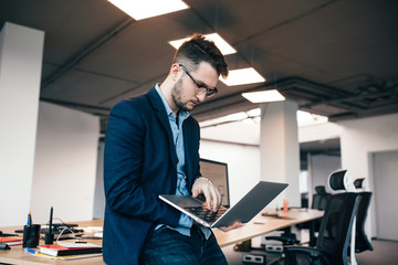 Serious  attractive man in glassess is standing near the workplace in office. He wears  blue shirt, dark jacket.  He is typing on laptop