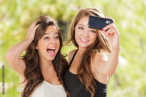 Fotografie, Obraz Two Attractive Mixed Race Girlfriends Using Their Smart Cell Phones For Selfie O