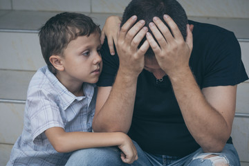 Portrait of young sad little boy and father sitting outdoors at the day time.