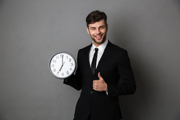 Poster - Young successful business man holding clock while showing thumb up gesture and looking at camera