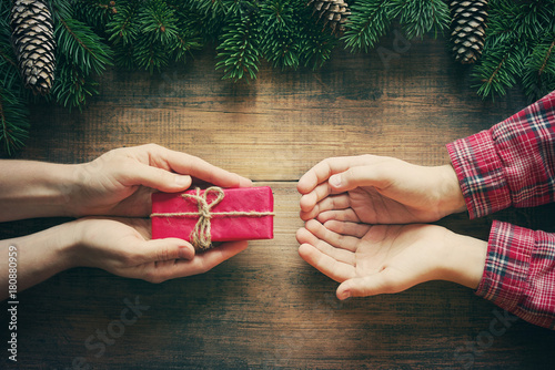 Fotografie, Obraz Red gift box in female hands and the child's hands accepting a gift, christmas fir tree on wooden background