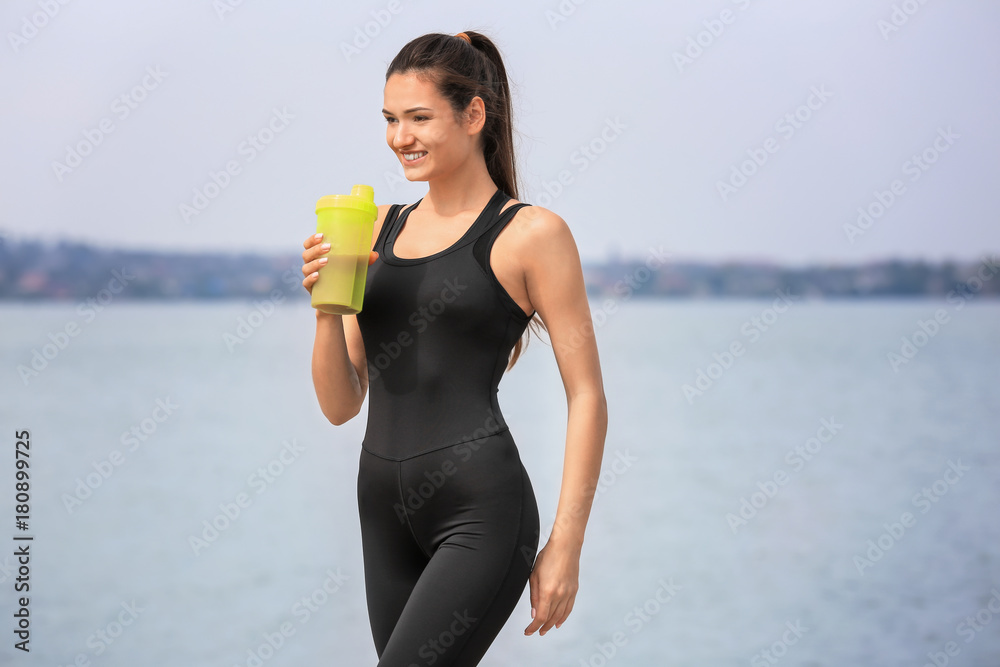 Beautiful young woman with protein shake on river bank