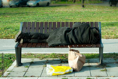 Fotografie, Obraz Homeless refugee man in black coat sleeps on the bench on the street