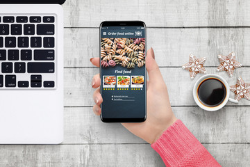 Female hand holding phone with food ordering app on white wooden desk surrounded with laptop and coffee